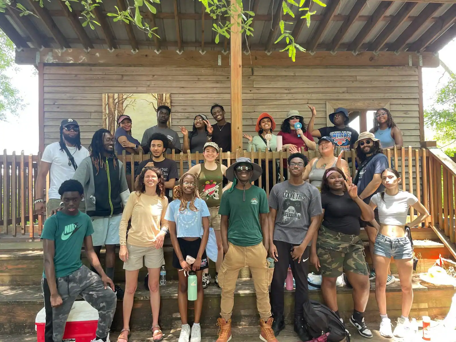 A group of people posing on wooden steps in front of a building, with trees in the background. Some are standing, and a few are seated, with various casual outfits and expressions.