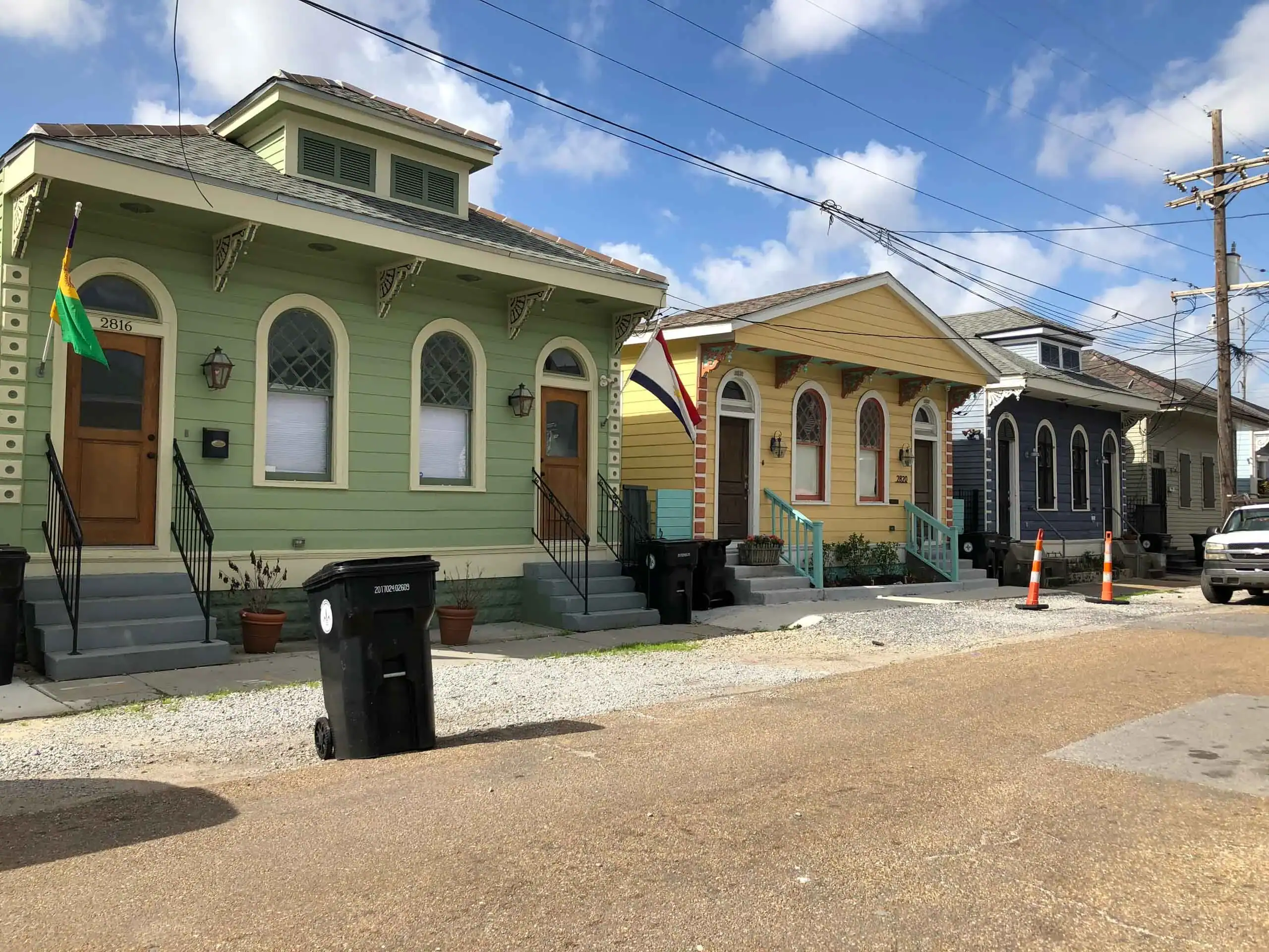 A row of colorful houses with arched windows and front steps line a street with utility poles and a parked truck under a clear blue sky. Trash bins and a traffic cone are on the sidewalk.