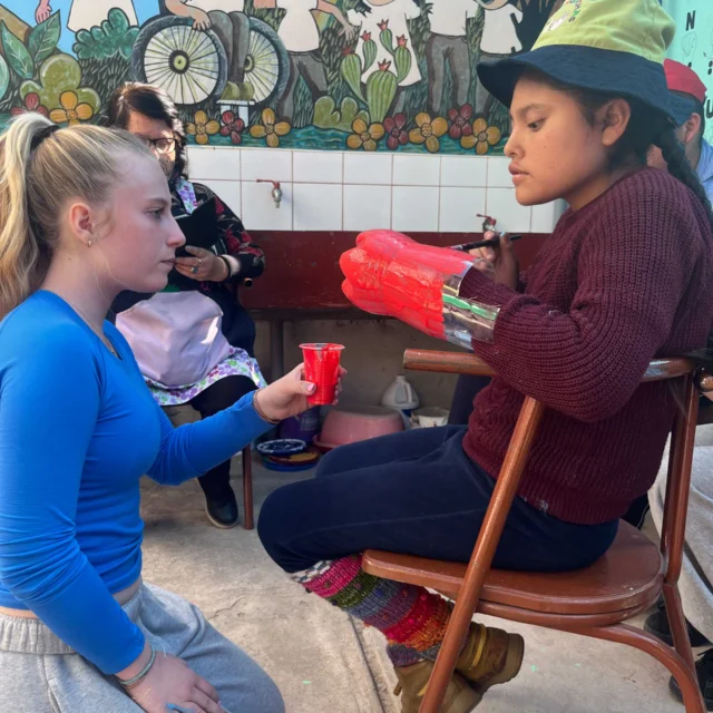 Two children engaged in craft activity; the child in the chair holds papier-mâché gloves, while the other kneels, holding a red brush. Colorful mural in the background.