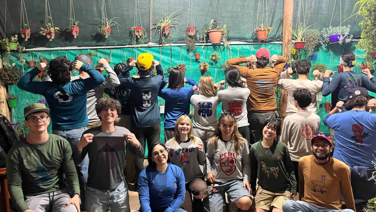 A group of people pose indoors, some smiling at the camera while others show the back of their shirts. Plants hang on the wall behind them.