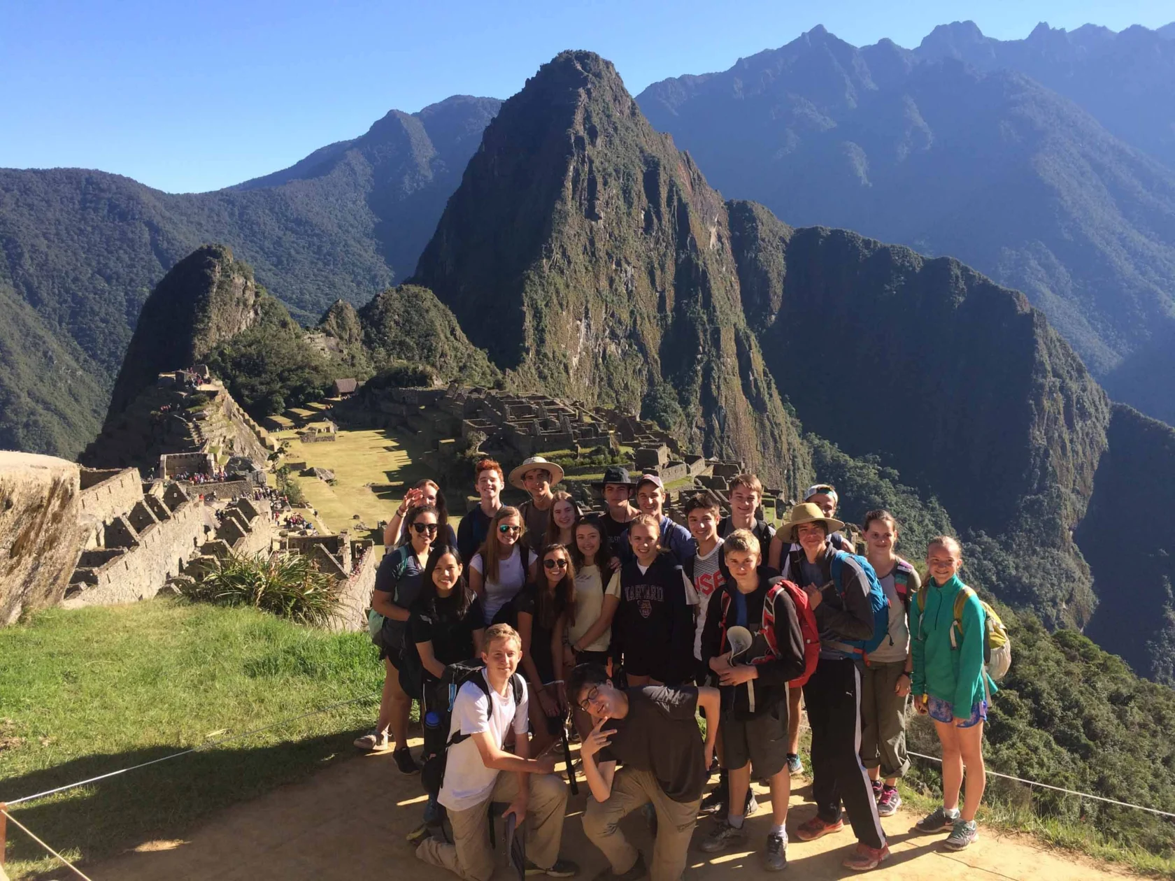 A group of people pose for a photo at the ancient Incan site of Machu Picchu, with the iconic mountain and ruins visible in the background under a clear blue sky.