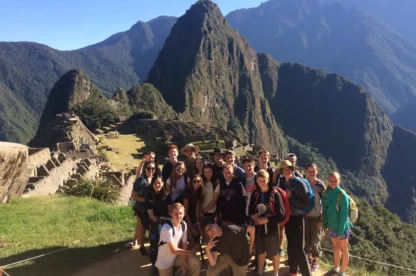 A group of people pose for a photo at the ancient Incan site of Machu Picchu, with the iconic mountain and ruins visible in the background under a clear blue sky.