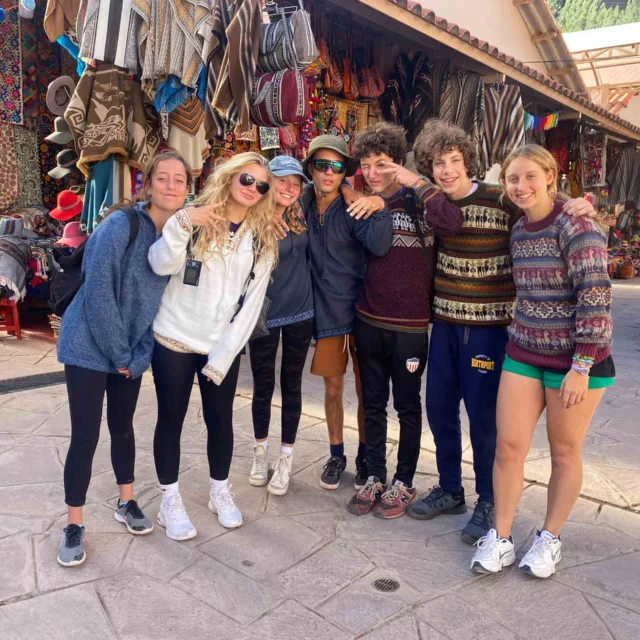 A group of seven young people in casual clothing stand together smiling in a vibrant outdoor market with textile goods on display.