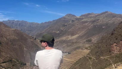 Person sitting on a hillside, wearing a white shirt and green cap, overlooking a valley with mountains and terraces under a clear blue sky.