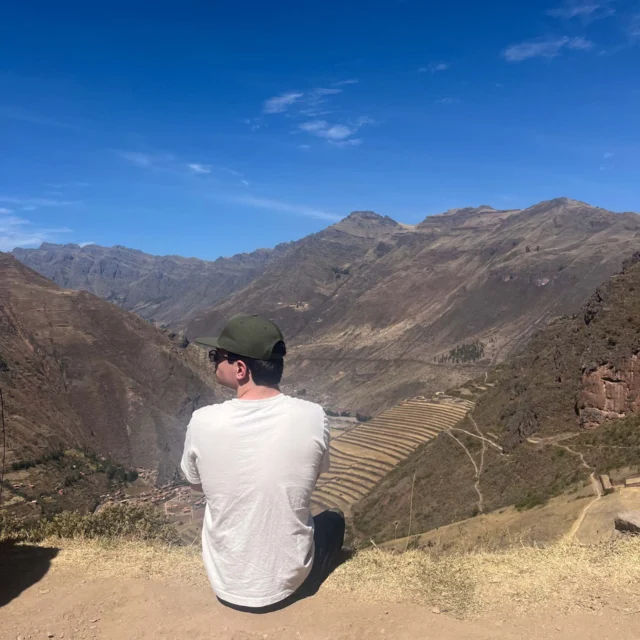 Person sitting on a hillside, wearing a white shirt and green cap, overlooking a valley with mountains and terraces under a clear blue sky.