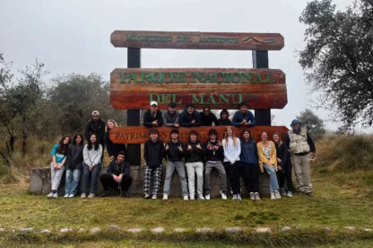 A group of people poses in front of a large wooden sign for Parque Nacional del Manu. The background is misty with trees and grass.