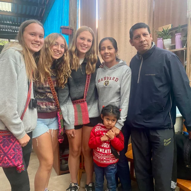 A group of six people, including four young women, one older woman, and a man, are posing together indoors. A child stands in front, wearing a red sweater.