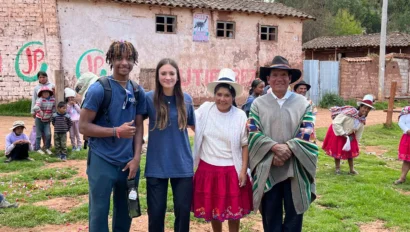 A group of people stands together outdoors in a rural area with an old building in the background. Children are playing nearby.