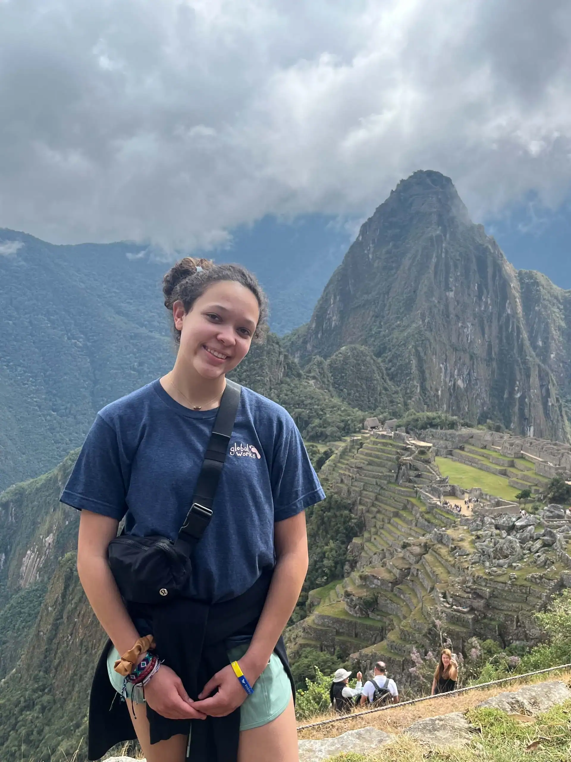 A young woman on a Peru student travel adventure stands in front of the ancient Incan city of Machu Picchu, with terraced ruins and a mountain peak visible in the background under cloudy skies.