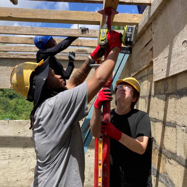 Three construction workers wearing helmets and gloves are working together to secure wooden beams on a building site under a partly cloudy sky.