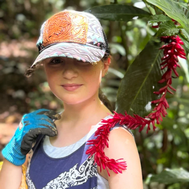 A person in a colorful cap stands near red tropical flowers in a lush, green forest, wearing a patterned shirt and a blue glove.