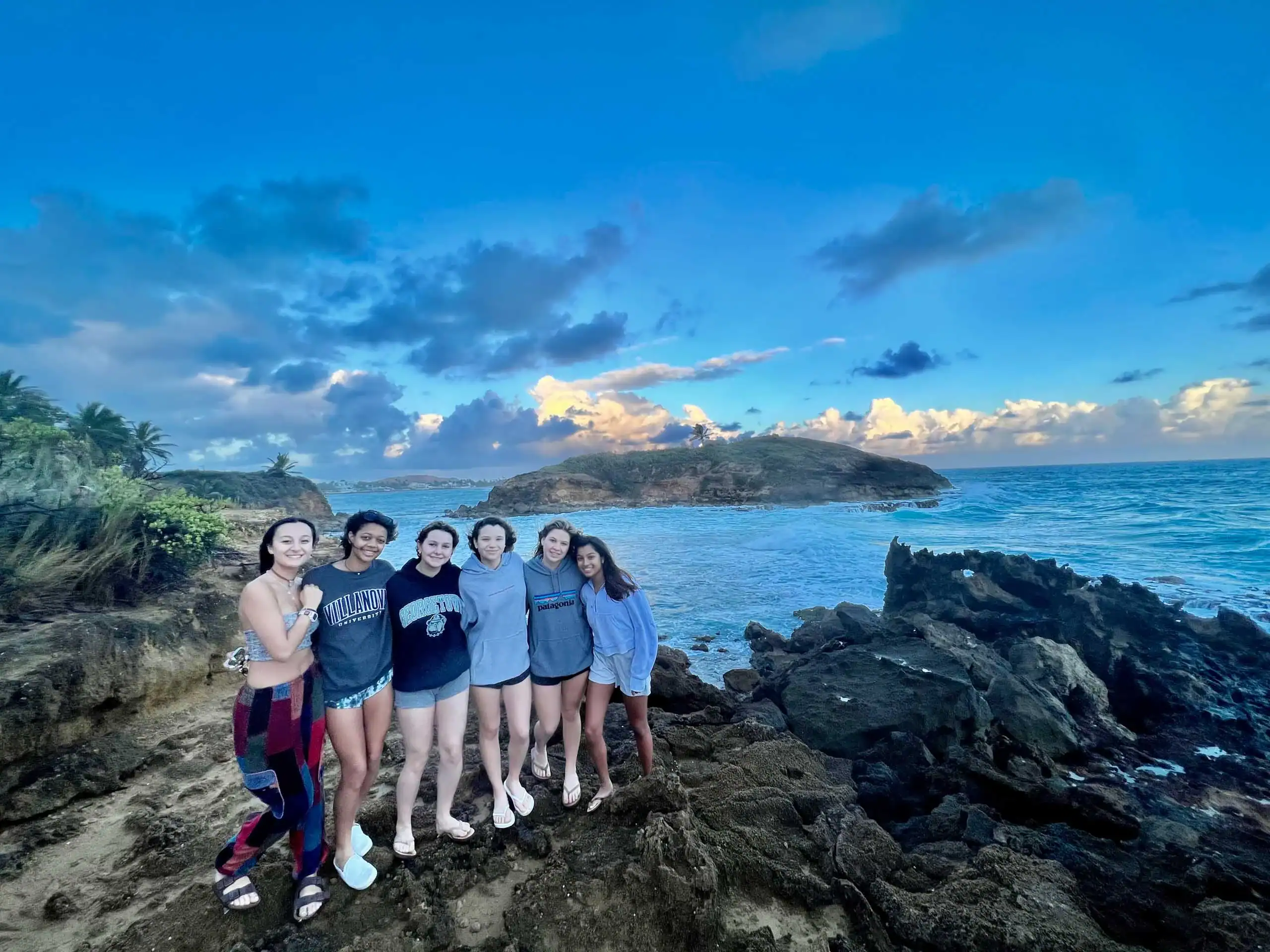 Six people stand together on rocky terrain by the ocean under a partly cloudy sky.