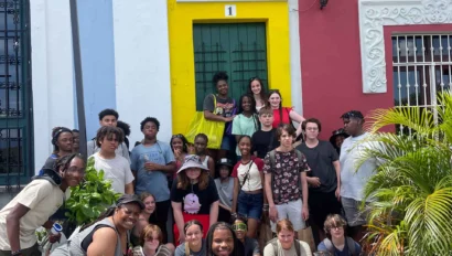 A diverse group of people poses in front of colorful buildings under bright sunlight. Some hold plants, and everyone appears relaxed and happy.