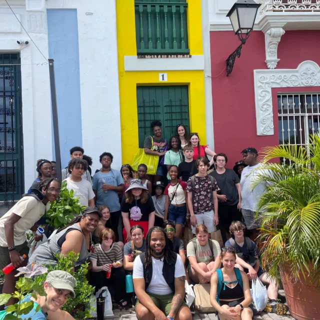 A diverse group of people poses in front of colorful buildings under bright sunlight. Some hold plants, and everyone appears relaxed and happy.