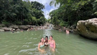 Three people stand in a natural green water pool, surrounded by trees and rocks, with others swimming in the background under a cloudy sky.