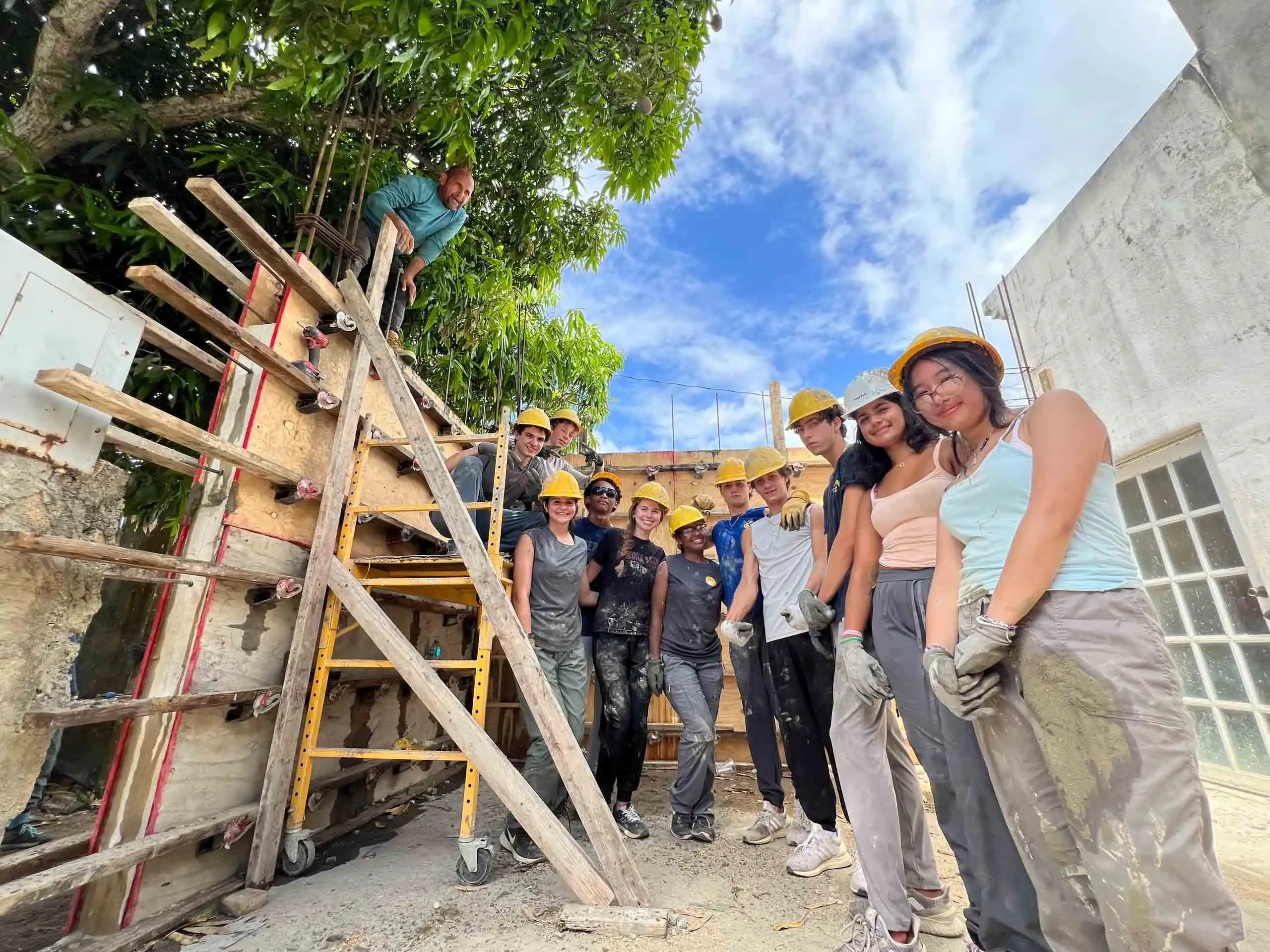 A group of teens yellow hard hats and work clothes pose at a construction site during a Puerto Rico hurricane relief service trip for teens, standing near a ladder and a partially built wall under a tree and blue sky.