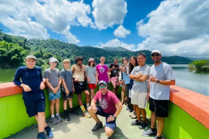 A group of people stands on a colorful bridge with a scenic lake and lush mountains in the background. They are smiling, and the weather is sunny with scattered clouds.