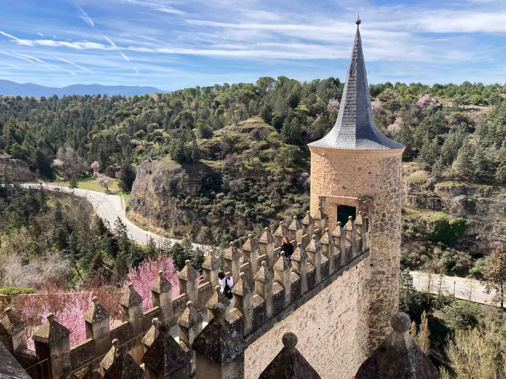 A medieval stone tower with a conical roof overlooks a lush, hilly landscape. People are visible on a walkway.