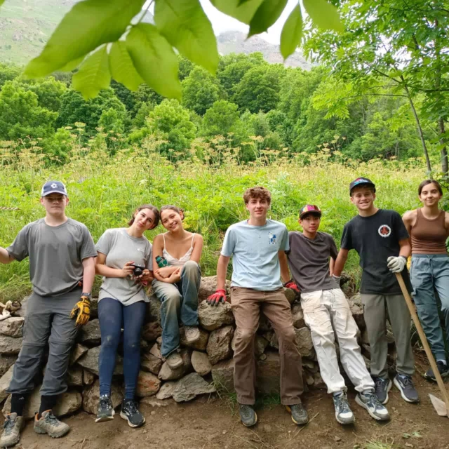 Seven young people in outdoor work clothes sit and stand by a low stone wall in a green, wooded area, with tools and gloves visible—capturing the spirit of a Costa Rica student travel adventure.