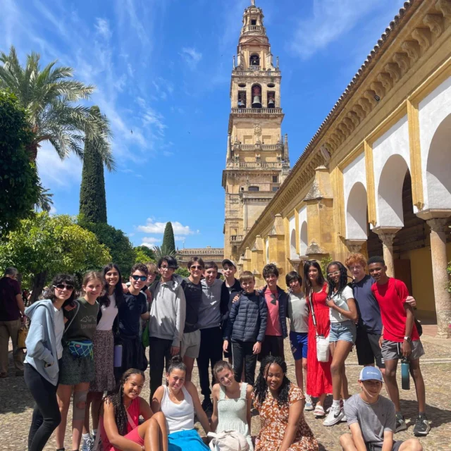 A group of people pose for a photo in front of a historic bell tower and arches, with a blue sky and palm tree in the background.