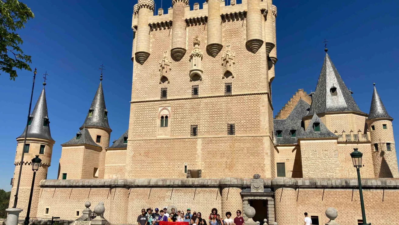 Group of people holding a Spanish flag in front of a historic castle with tall, ornate towers and a clear blue sky.