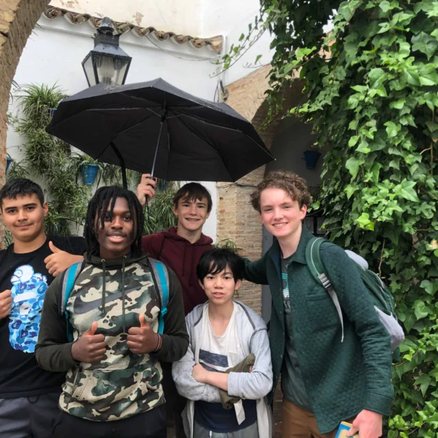 Five people standing in front of a brick archway with greenery. One holds an umbrella. Some are smiling and giving a thumbs up.