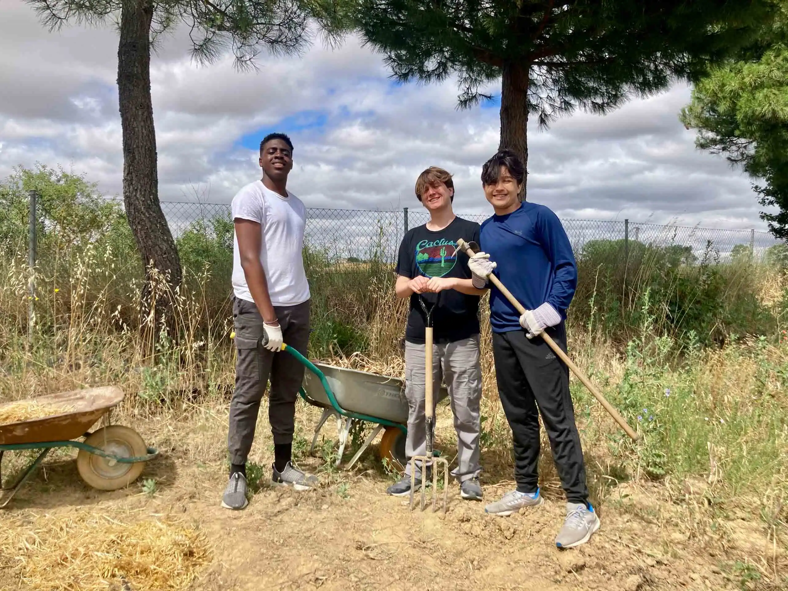 Three students stand outdoors on dry ground, holding gardening tools and wearing gloves, with a wheelbarrow and tall grass behind them—capturing a moment from a Spain school trip under a partly cloudy sky.