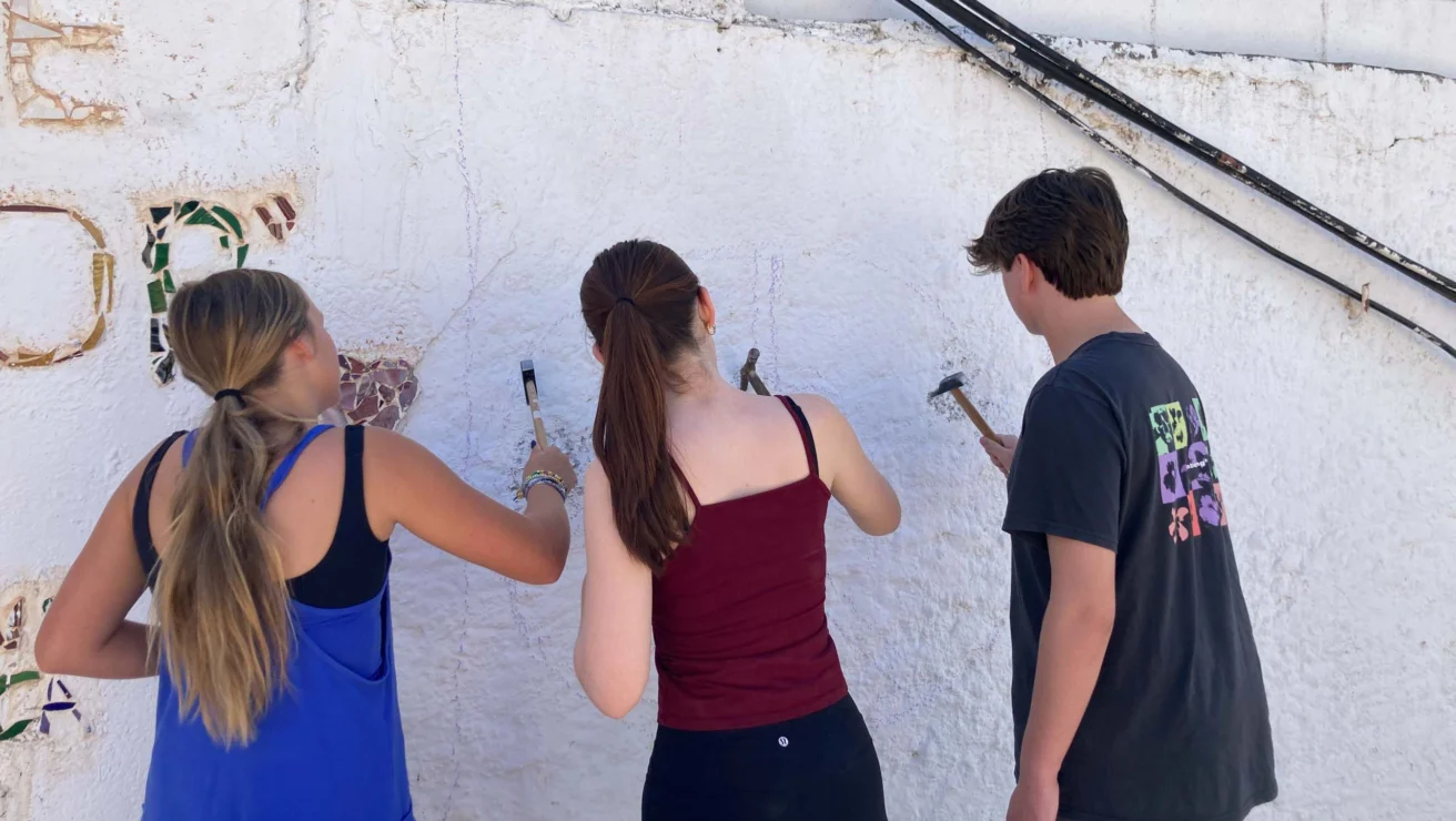 Three people are painting a white wall. Two are holding brushes, one is observing. The setting appears to be outdoors.