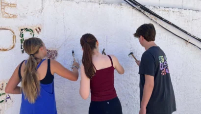 Three people are painting a white wall. Two are holding brushes, one is observing. The setting appears to be outdoors.