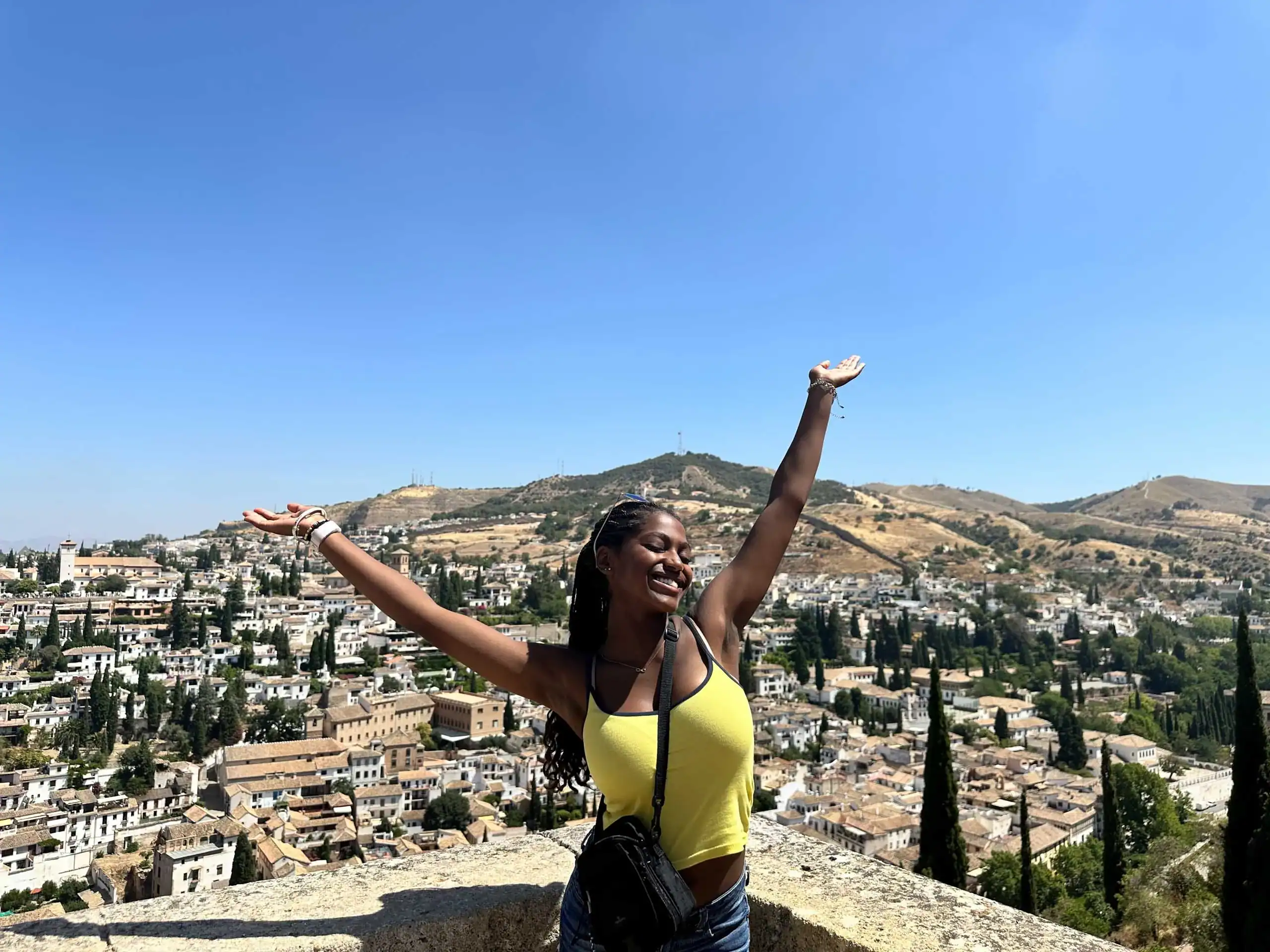 Student in a yellow top stands with arms raised, smiling, enjoying a sunny hillside town and clear blue sky during her Spain school trip.