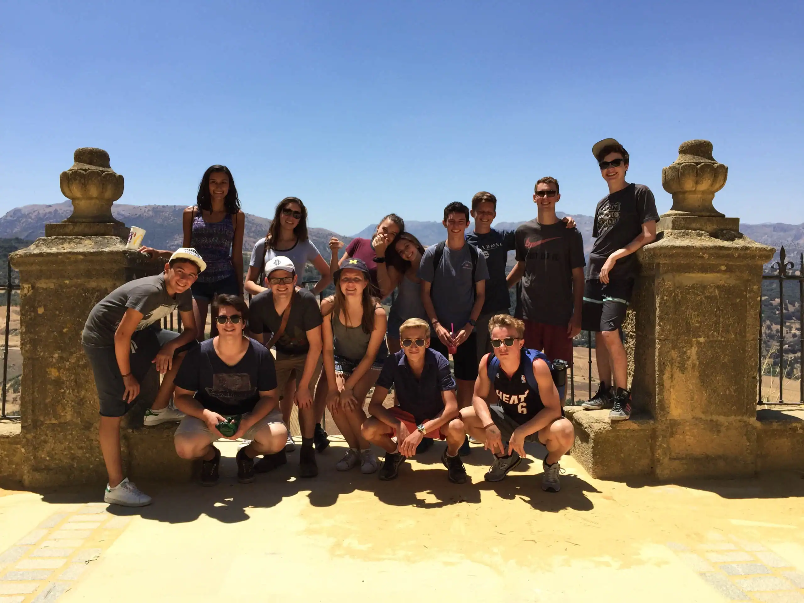 A group of thirteen people from the Spain summer travel program poses together outdoors on a sunny day, standing and kneeling in front of a stone railing with a scenic background.
