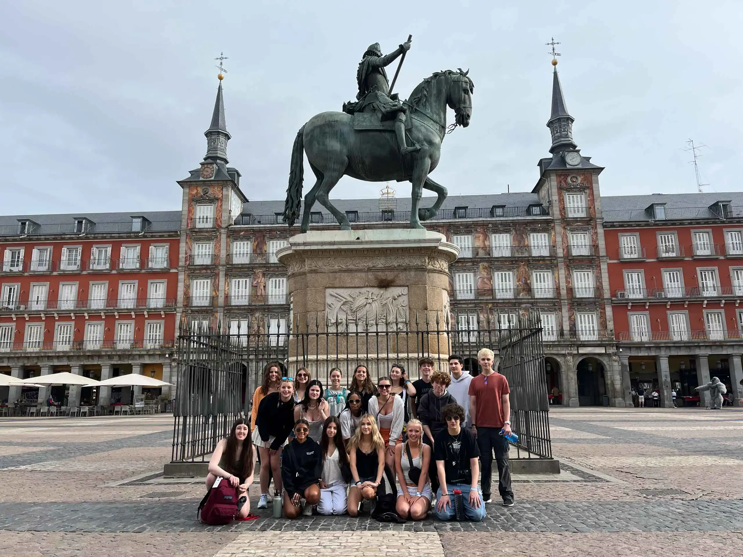 A group of young people in the Spain summer travel program pose in front of an equestrian statue in the center of a large plaza with historic buildings in the background.