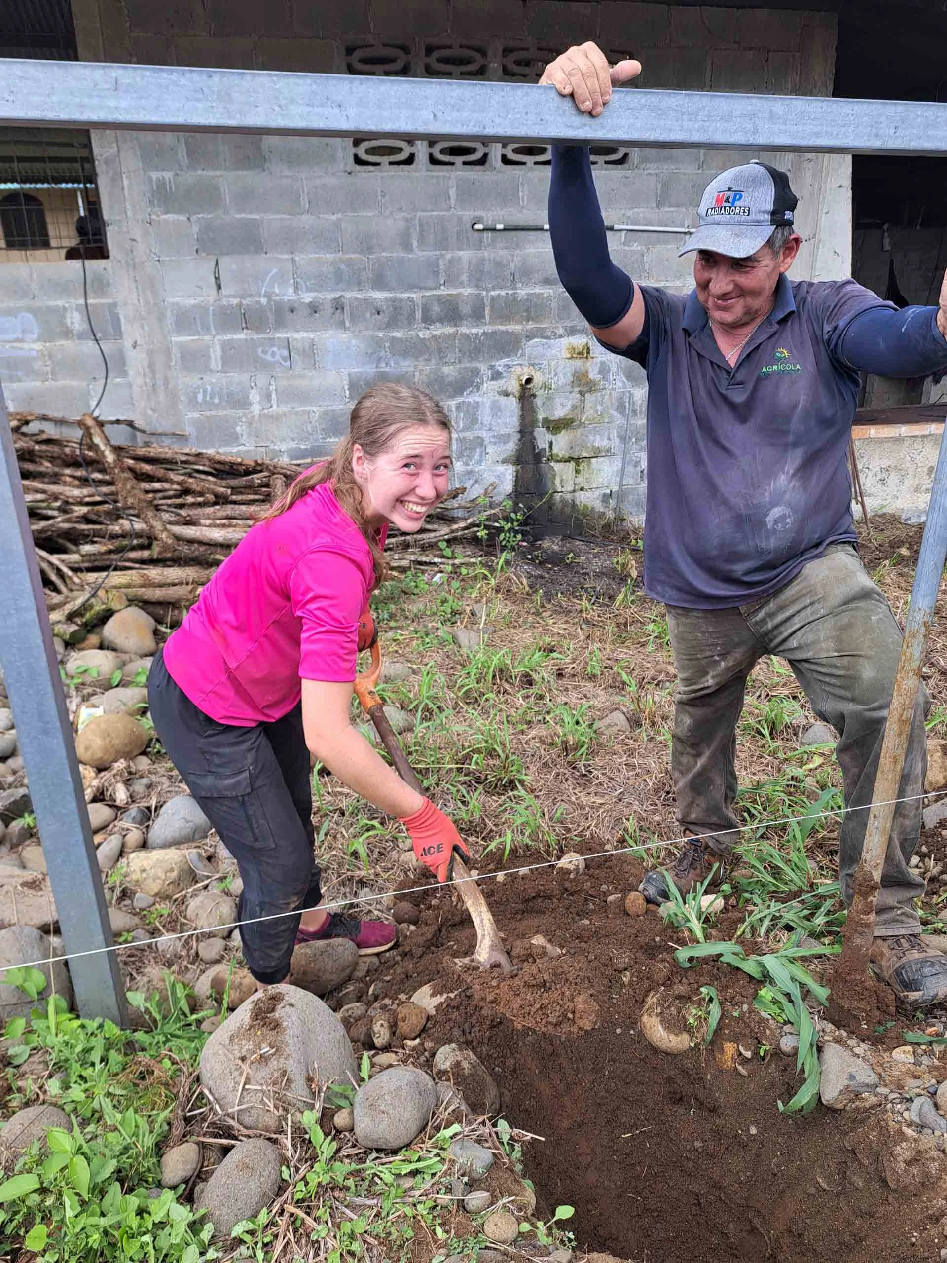 A high school student and a man work together outdoors, digging in the dirt near a building with rocks, plants, and a pile of branches—possibly as part of a Spanish immersion trip focused on hands-on learning.