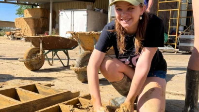 Person kneeling on the ground working with mud in wooden molds. A wheelbarrow and building materials are in the background. Outside, sunny day.