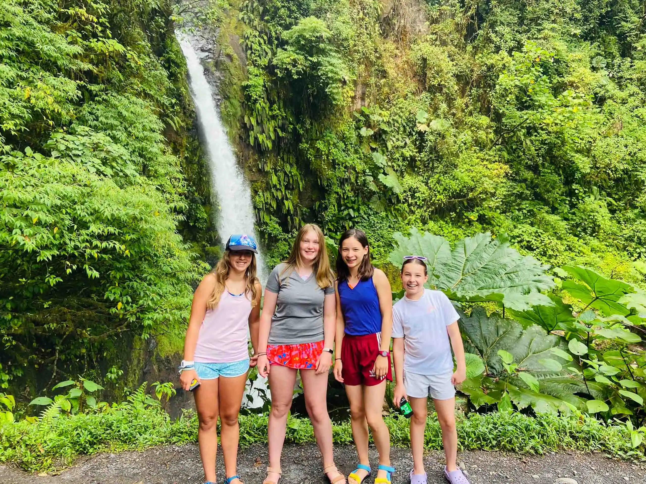 Four girls, during student travel to Costa Rica, stand on a path in front of a tall waterfall surrounded by dense green vegetation.