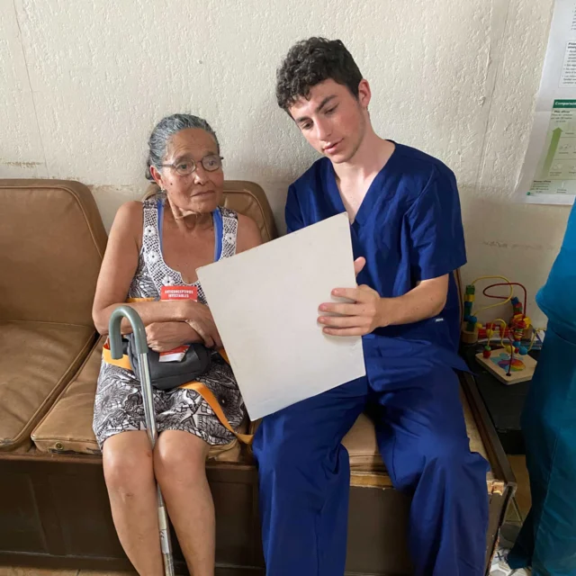 A Global Works student in blue scrubs sits beside an older woman with a cane, showing her a document while they sit on a bench indoors—part of a Costa Rica student travel program focused on compassionate senior care.