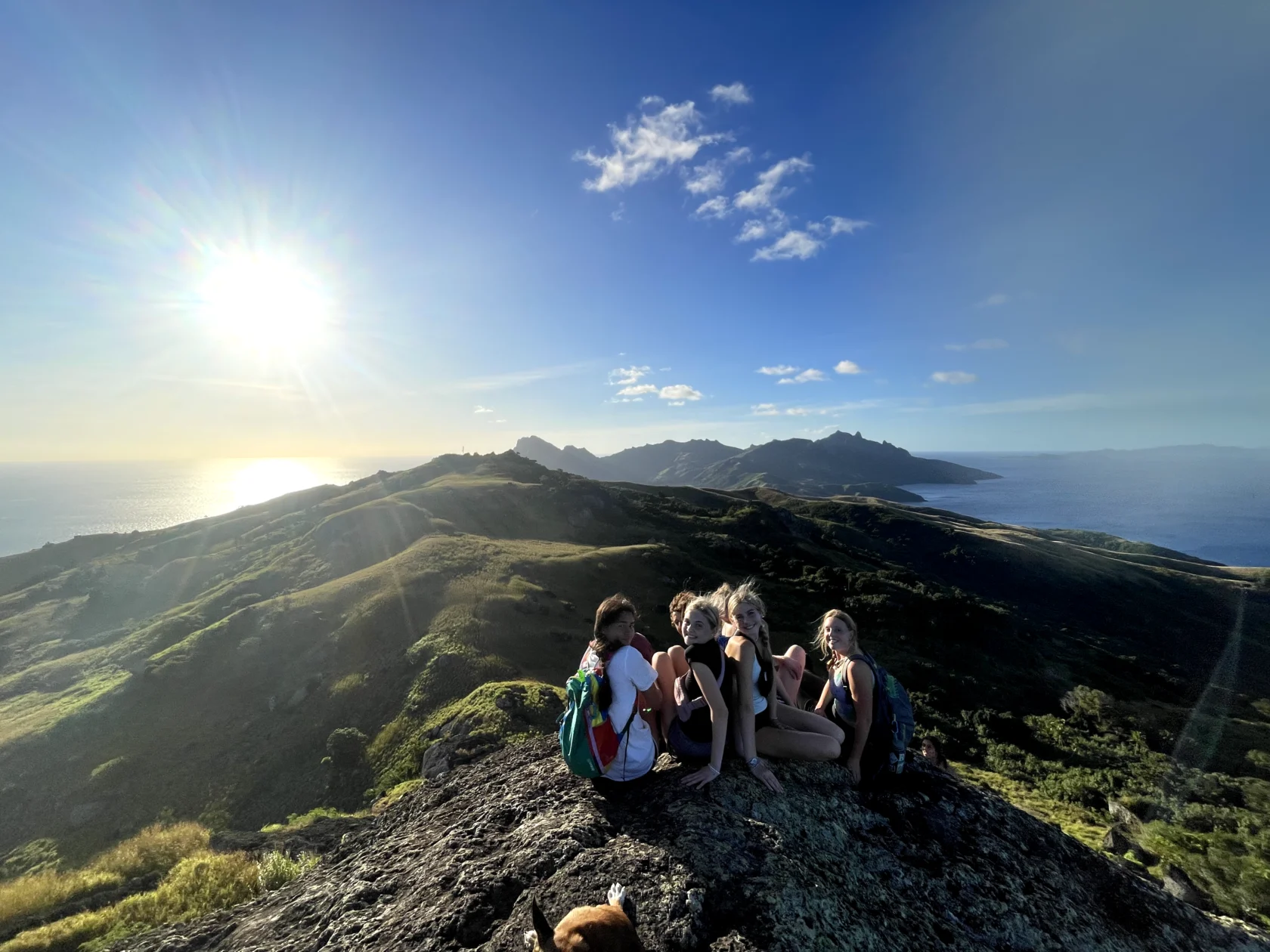 Four people sit together on a rocky hilltop, overlooking a scenic landscape with the sun shining brightly in the sky.