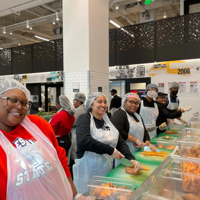 A group of people wearing hairnets and aprons prepare food at a counter in a bright kitchen space.
