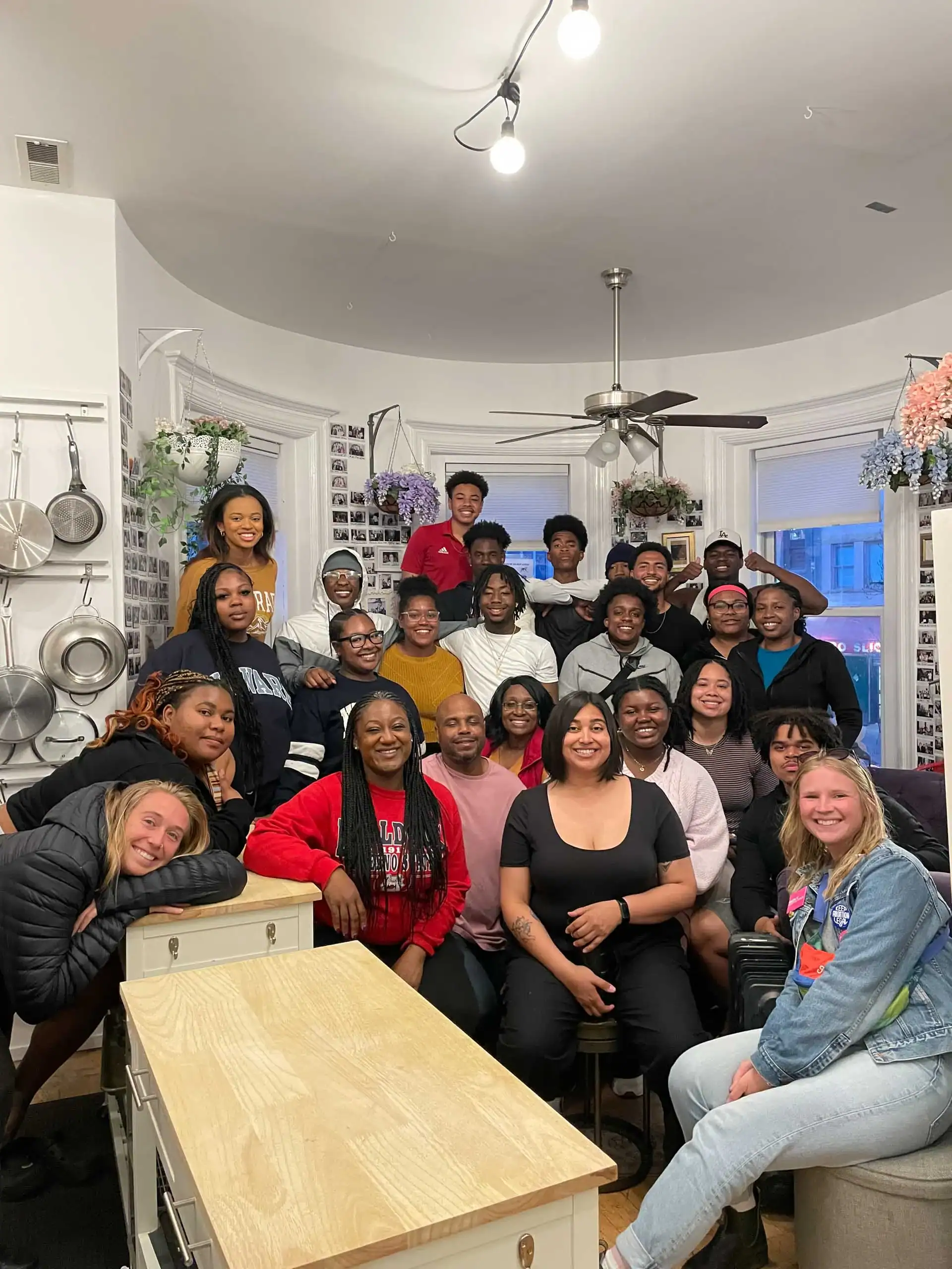 A group of people gathered in a cozy kitchen, smiling at the camera. Pots and pans hang on the wall.