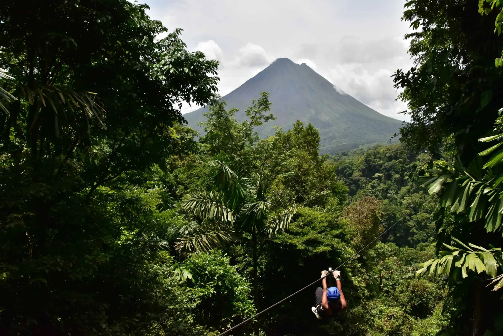 A student ziplining through a dense green forest, with a distant volcano in the background under a partly cloudy sky—an unforgettable adventure perfect for any school travel program by Global Works.