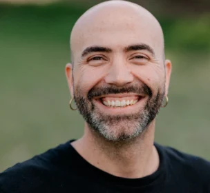 A man with a beard and hoop earrings smiles broadly. He is wearing a black Volcom Stone T-shirt. A blurred green background is visible.