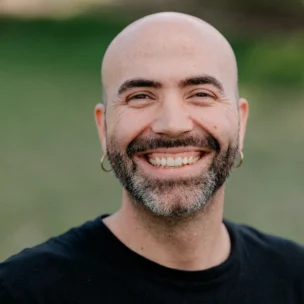 A man with a beard and hoop earrings smiles broadly. He is wearing a black Volcom Stone T-shirt. A blurred green background is visible.
