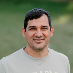 A man with short dark hair stands outdoors wearing a gray t-shirt with "A. EAGLE USA" on the front, smiling at the camera.