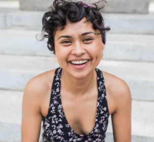 A person with curly hair and a headband smiles while sitting on outdoor steps, wearing a floral-patterned top.