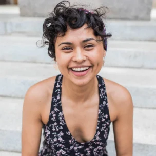 A person with curly hair and a headband smiles while sitting on outdoor steps, wearing a floral-patterned top.