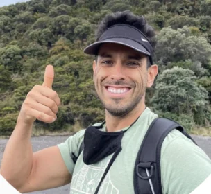 A person outdoors, smiling and giving a thumbs-up, wearing a black visor and backpack, with a green forest background.