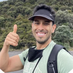 A person outdoors, smiling and giving a thumbs-up, wearing a black visor and backpack, with a green forest background.