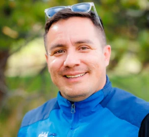 A person wearing a blue jacket and sunglasses on their head smiles outdoors with trees in the background.