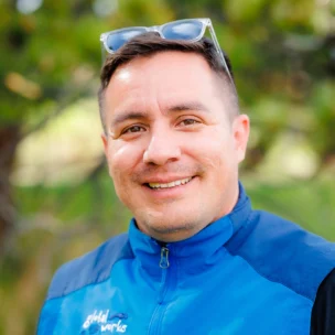 A person wearing a blue jacket and sunglasses on their head smiles outdoors with trees in the background.