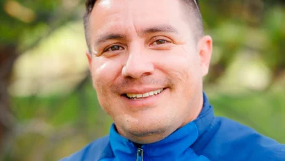 A person wearing a blue jacket and sunglasses on their head smiles outdoors with trees in the background.